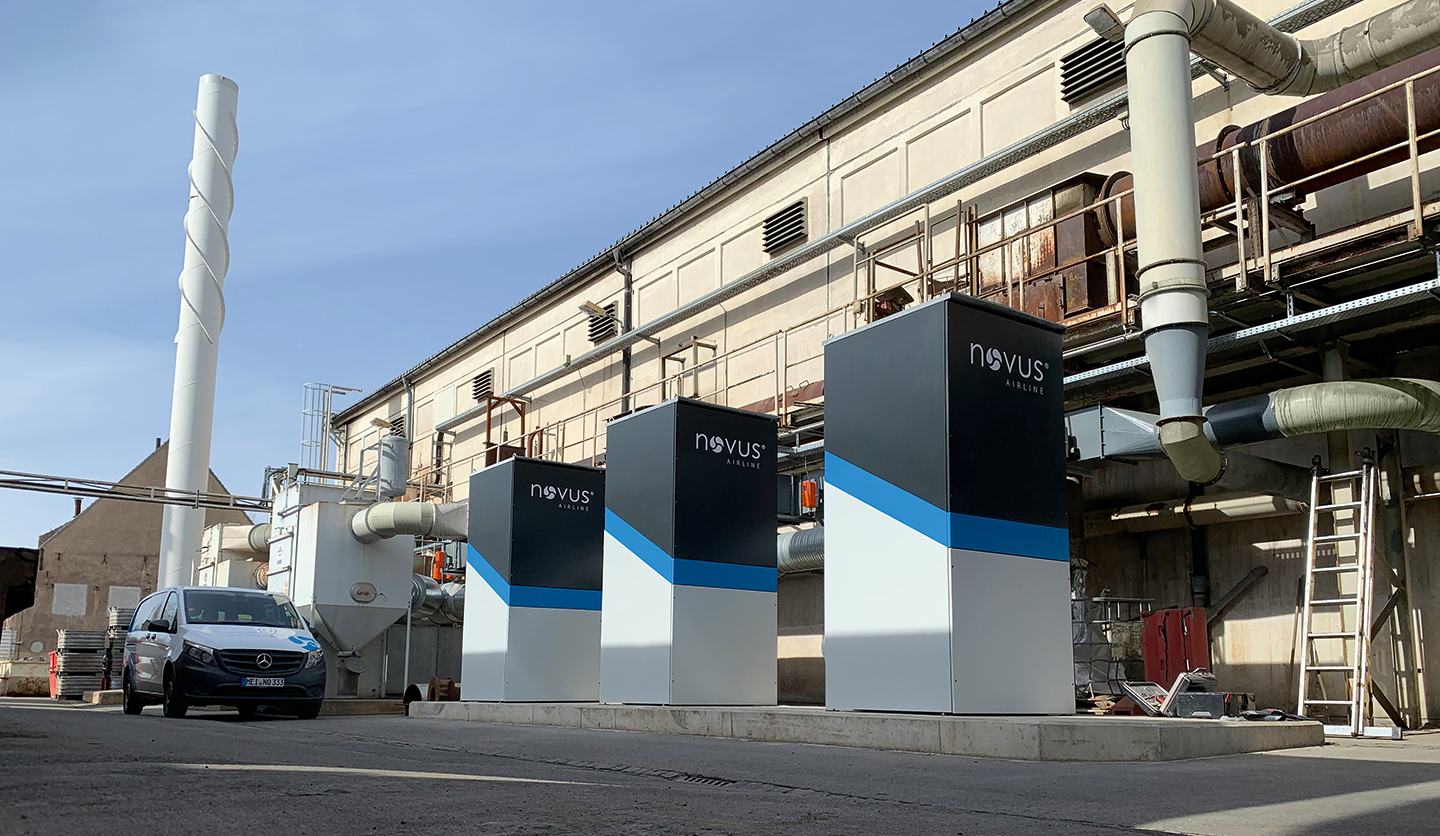Drei Absaug- und Filteranlagen vom Typ ‚Novus Airline‘ vor einer Industriehalle mit Rohrleitungen und einem Firmenfahrzeug im Vordergrund.