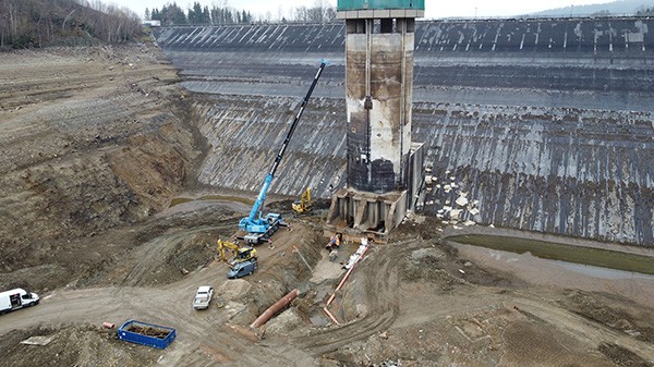 Bauarbeiten an einer teilweise entleerten Talsperre mit Kran, Baumaschinen und einem zentralen Turmbauwerk